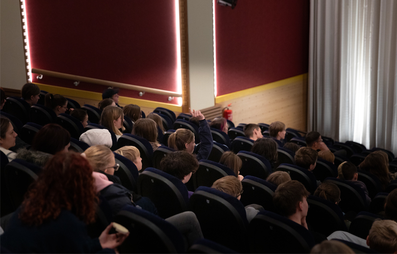 Abgebildet ist ein Kinosaal aus der Perspektive von hinten. Schüler und Schülerinnen sitzen in den Reihen. Ein Mensch meldet sich, um eine Frage zu stellen.