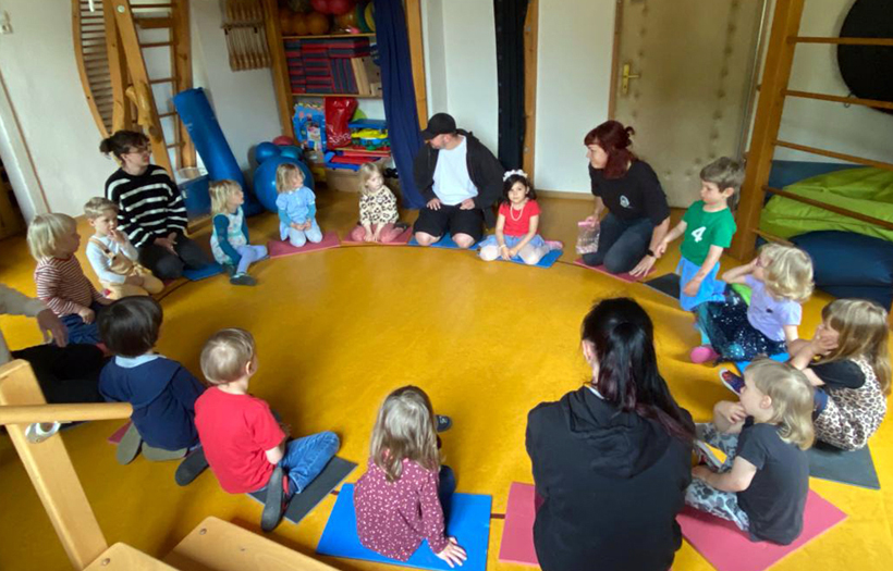 In einem Zimmer der Kita sitzen Erwachsene und Kinder in einem Kreis auf dem Boden. Sie sprechen gemeinsam über das Projekt des Eisvogels.