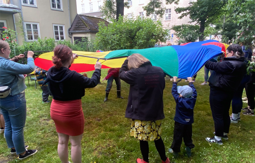 In einem Garten stehen Kinder und Erwachsene im Kreis. Sie alle halten zusammen ein großes Tuch in Regenbogenfarben.